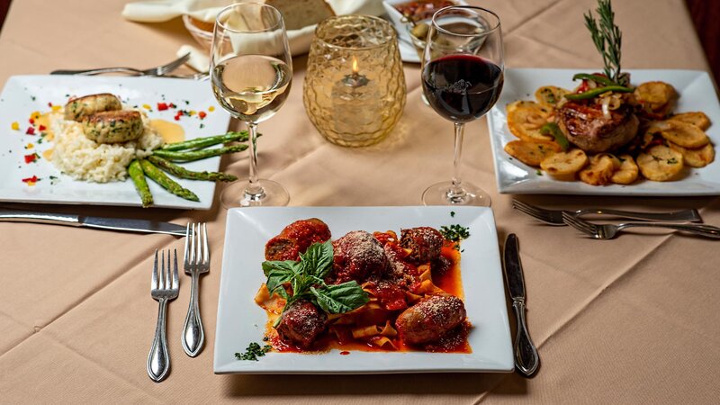 Three plated entrees with focus on Marco's Mothers Sunday Sauce entree with red and white wine in glasses